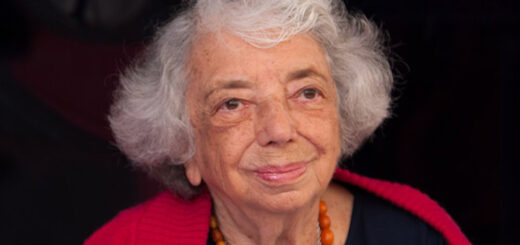 Porträt der Holocaust-Überlebenden und Zeitzeugin Margot Friedländer, lächelnd mit weißem Haar, rotem Schal und orangefarbener Bernsteinkette.
