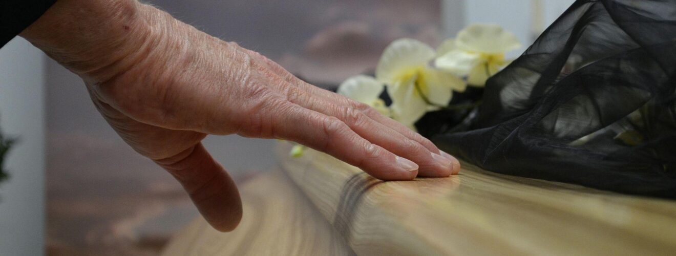Nahaufnahme einer Hand, die behutsam über die glatte Holzoberfläche eines Sarges streicht; daneben liegen helle Blumen und ein schwarzes Tuch – ein stiller Moment des Abschieds bei einer Trauerfeier.