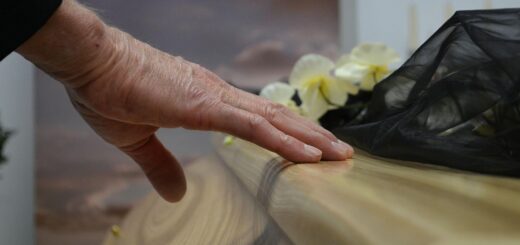 Nahaufnahme einer Hand, die behutsam über die glatte Holzoberfläche eines Sarges streicht; daneben liegen helle Blumen und ein schwarzes Tuch – ein stiller Moment des Abschieds bei einer Trauerfeier.
