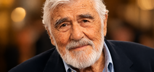 Porträt des älteren Schauspielers Mario Adorf mit weißem Haar und vollem Bart, warm beleuchtet vor unscharfem Hintergrund. Er trägt ein dunkles Sakko und ein hellblaues Hemd, blickt ruhig in die Kamera und wirkt würdevoll, freundlich und nachdenklich.